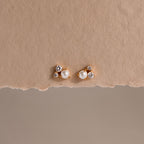 Pearl Diamond Cluster Studs in 18K Gold with pearls and small crystals rest on a textured beige background—elegant stud earrings for everyday wear.