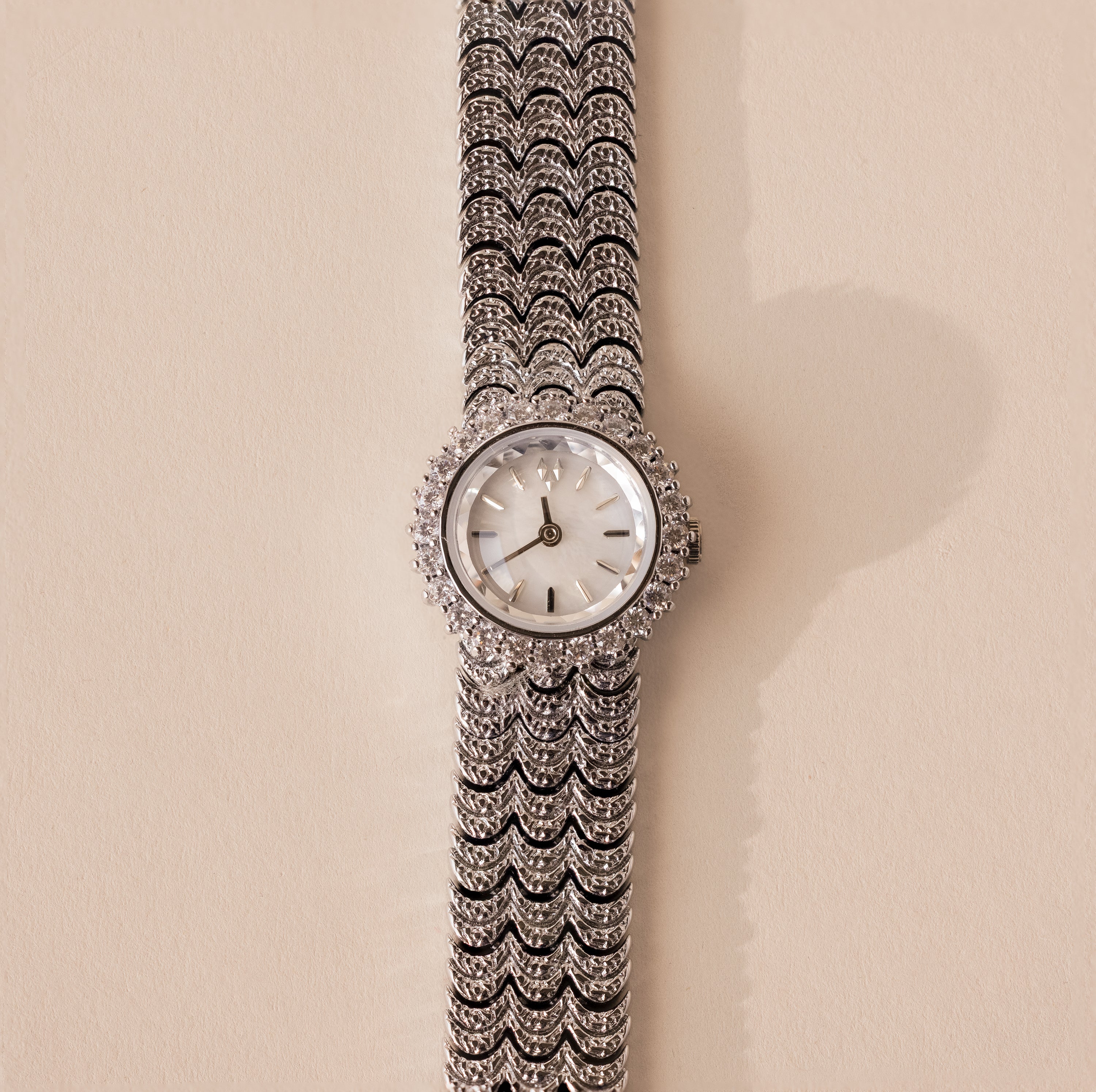 The Pave Scalloped Timepiece is a silver luxury wristwatch with a pavé bezel, diamond accents, and a patterned metal band, elegantly showcased on a beige background.