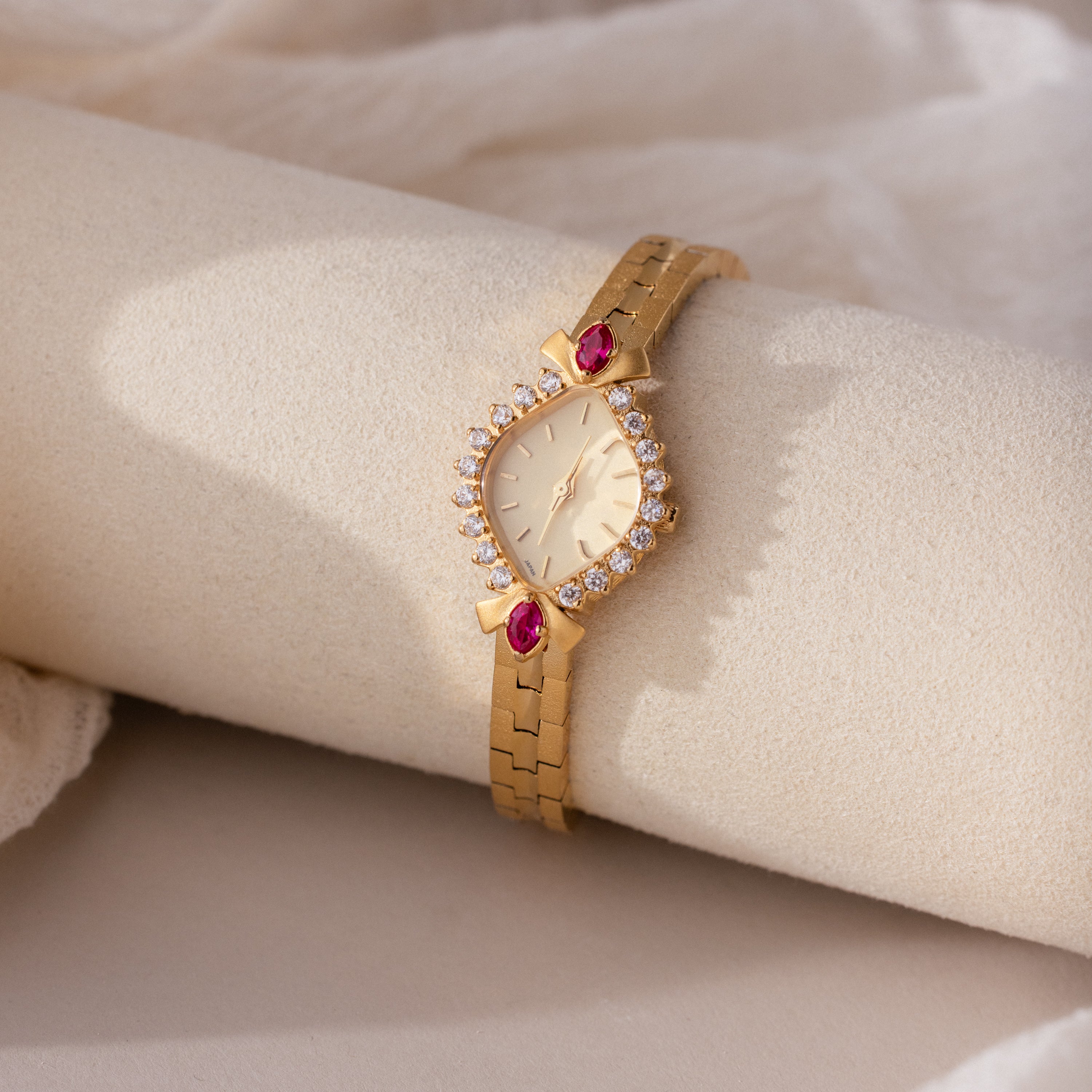 The Art Deco Ruby Timepiece features a gold bracelet, teardrop-shaped face, ruby accents, and diamond-like stones on soft beige fabric—capturing retro glamour for an elegant look.