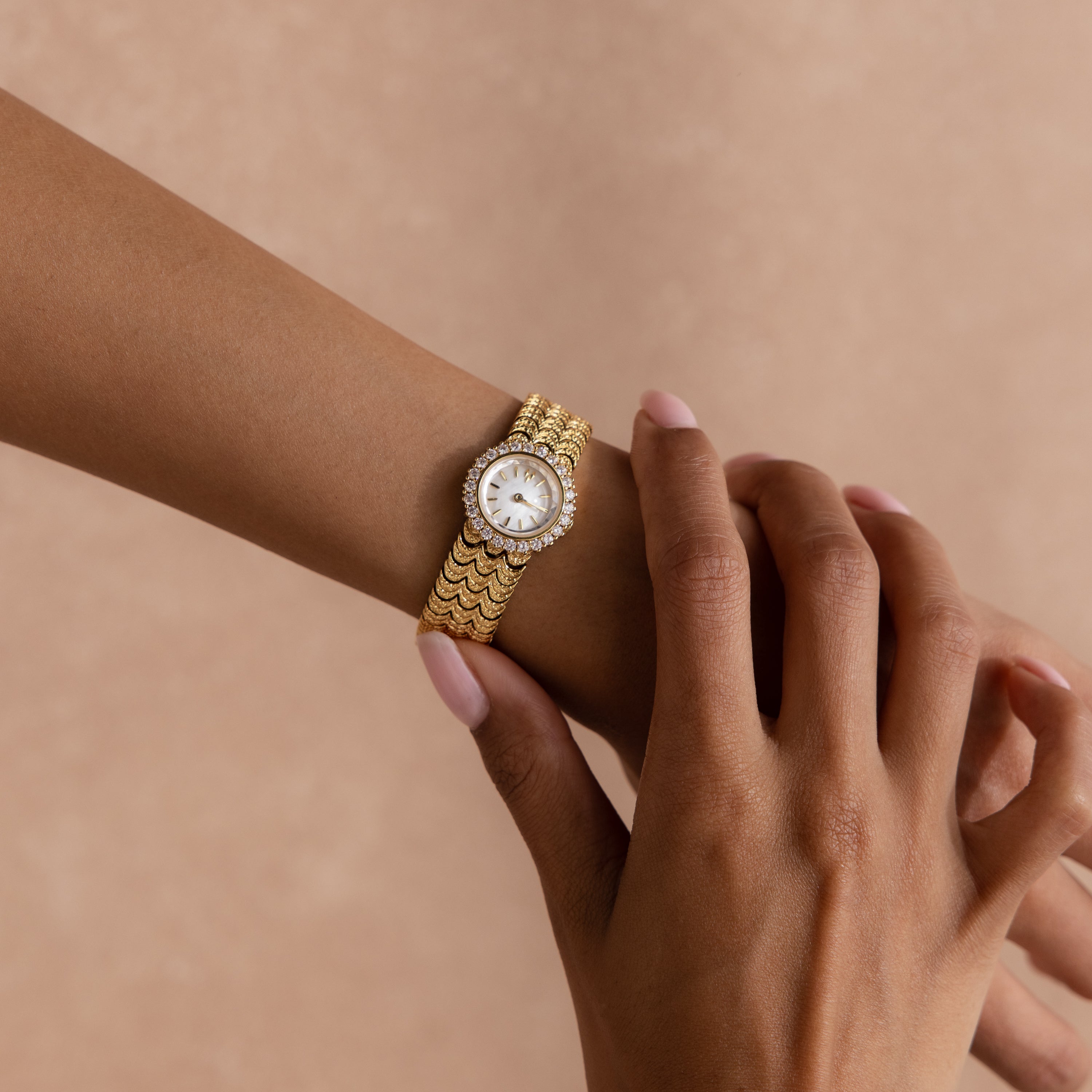 A hand wearing the Pave Scalloped Timepiece, a gold watch with a jeweled round face and pavé bezel, rests gracefully against a neutral background.