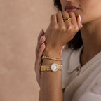 A woman in a cream blouse and gold jewelry rests her hand on her chin beside the Pave Scalloped Timepiece, a bracelet watch that adds an elegant touch.