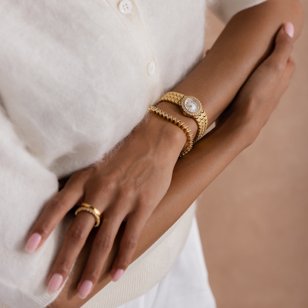 A woman in a white top crosses her arms, showcasing the Pave Scalloped Timepiece in 18K Gold, with its pavé bezel and scallop-link bracelet, paired with a gold ring.
