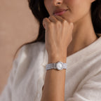 A woman in a white sweater showcases the Pave Scalloped Timepiece in White Gold with a scallop-link bracelet on her left wrist.