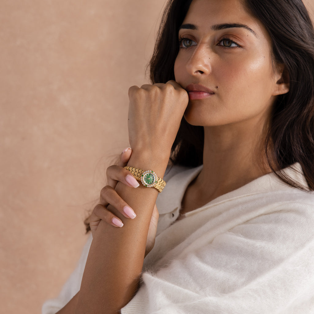 A woman in a white sweater rests her chin on her hand, wearing the Emerald Scalloped Timepiece—an elegant gold watch with an emerald-green dial and a scallop-link bracelet.