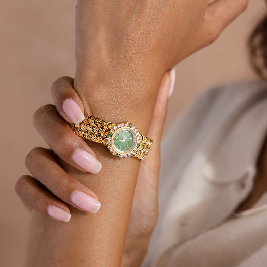 A hand with manicured nails wears the Emerald Scalloped Timepiece, showcasing a gold scallop-link bracelet and an emerald-green jeweled dial.