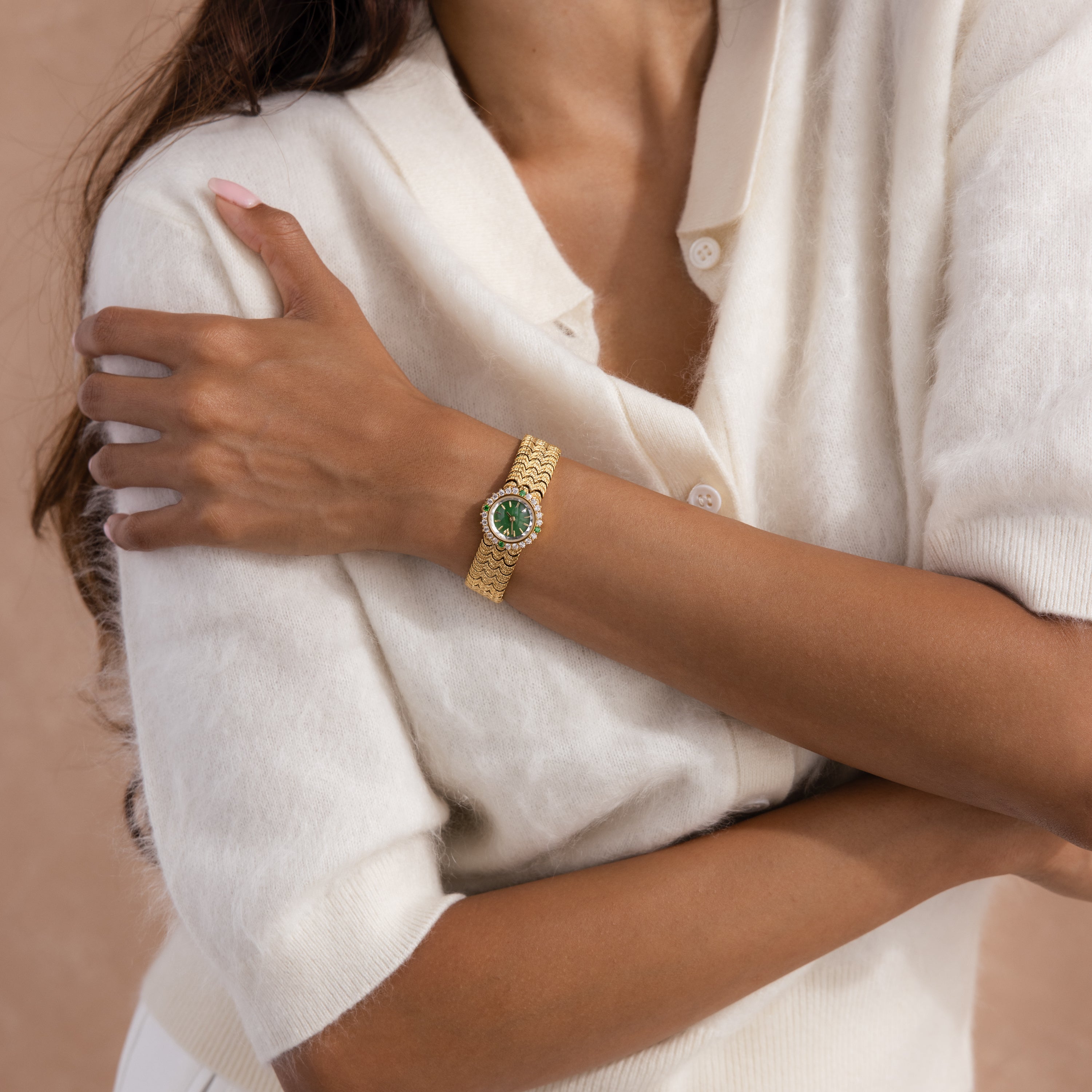 A woman in a cream sweater crosses her arms, displaying the Emerald Scalloped Timepiece featuring a scallop-link bracelet and an elegant emerald-green dial on her wrist.