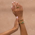 Two hands with light pink nails wear gold jewelry, including a scallop-link bracelet and the Emerald Scalloped Timepiece, both adorned with green stones, set against a beige background.