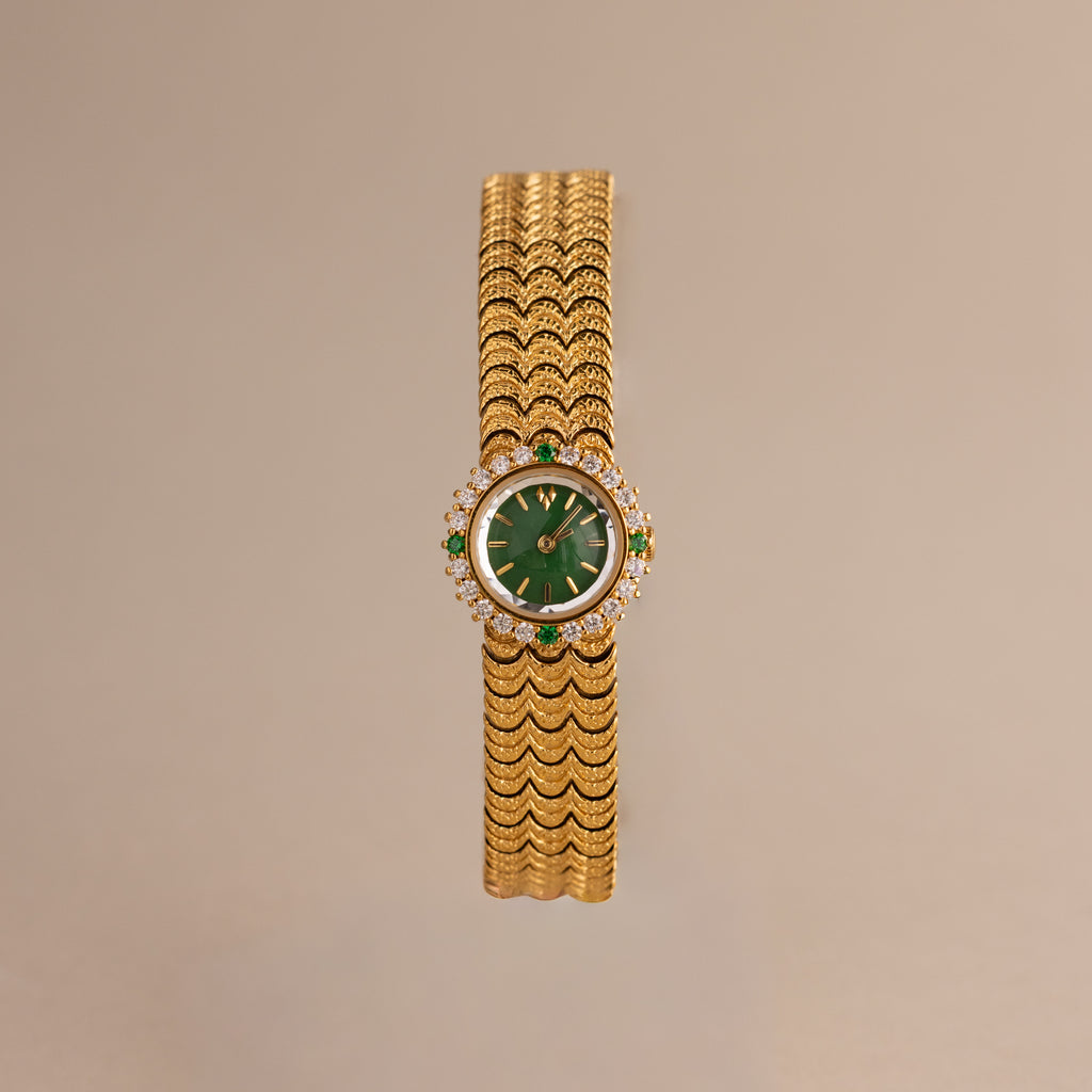 The Emerald Scalloped Timepiece showcases an emerald-green dial, diamond and emerald accents, and a scallop-link gold bracelet, displayed on a neutral background.