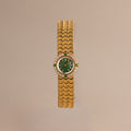 The Emerald Scalloped Timepiece showcases an emerald-green dial, diamond and emerald accents, and a scallop-link gold bracelet, displayed on a neutral background.