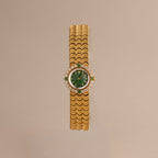 The Emerald Scalloped Timepiece showcases an emerald-green dial, diamond and emerald accents, and a scallop-link gold bracelet, displayed on a neutral background.