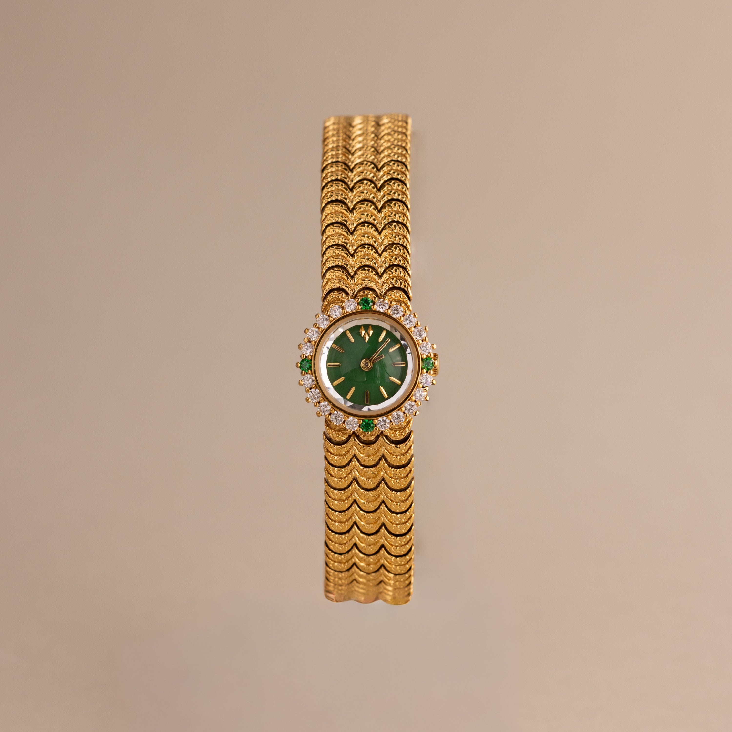 The Emerald Scalloped Timepiece showcases an emerald-green dial, diamond and emerald accents, and a scallop-link gold bracelet, displayed on a neutral background.