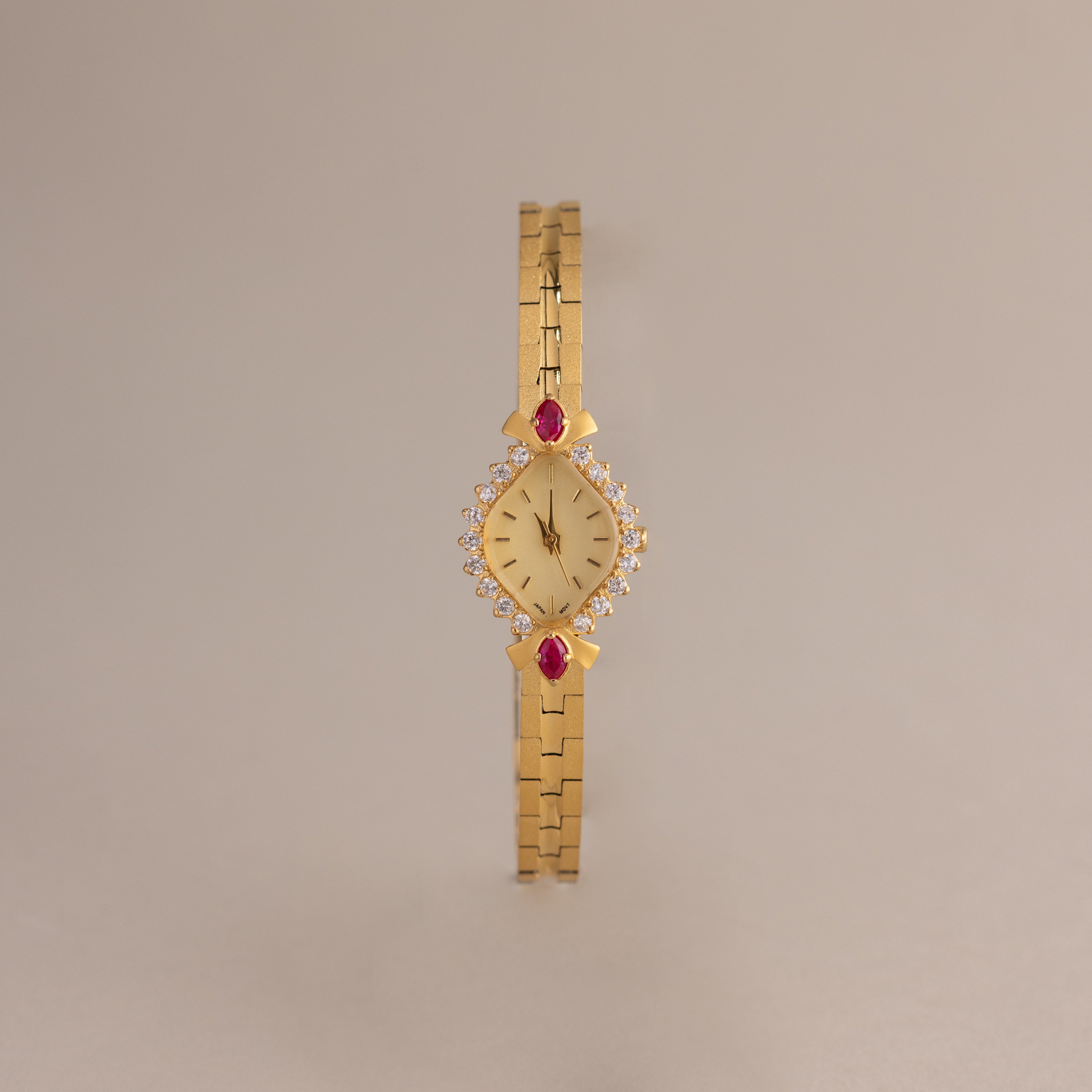 The Art Deco Ruby Timepiece features a gold bracelet and gemstone accents on the face, evoking retro glamour, shown against a plain beige background.