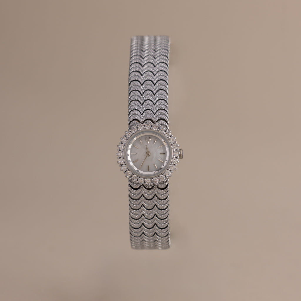 The Pave Scalloped Timepiece features a silver round face, scallop-link bracelet with diamond accents, and a pavé bezel, showcased on a beige background.