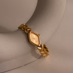 Product shot of a gold oval-faced watch with a chevron link bracelet, displayed draped over a curved ceramic object.