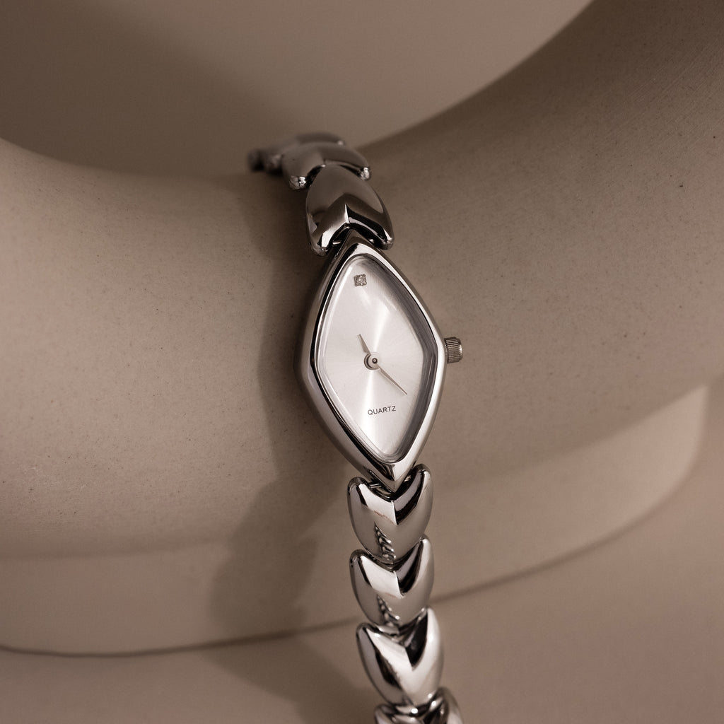 Angled view of a silver wristwatch with a marquise-shaped dial, resting against a neutral ceramic arc, showcasing the dial's shimmer and streamlined bracelet.