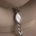 Angled view of a silver wristwatch with a marquise-shaped dial, resting against a neutral ceramic arc, showcasing the dial's shimmer and streamlined bracelet.