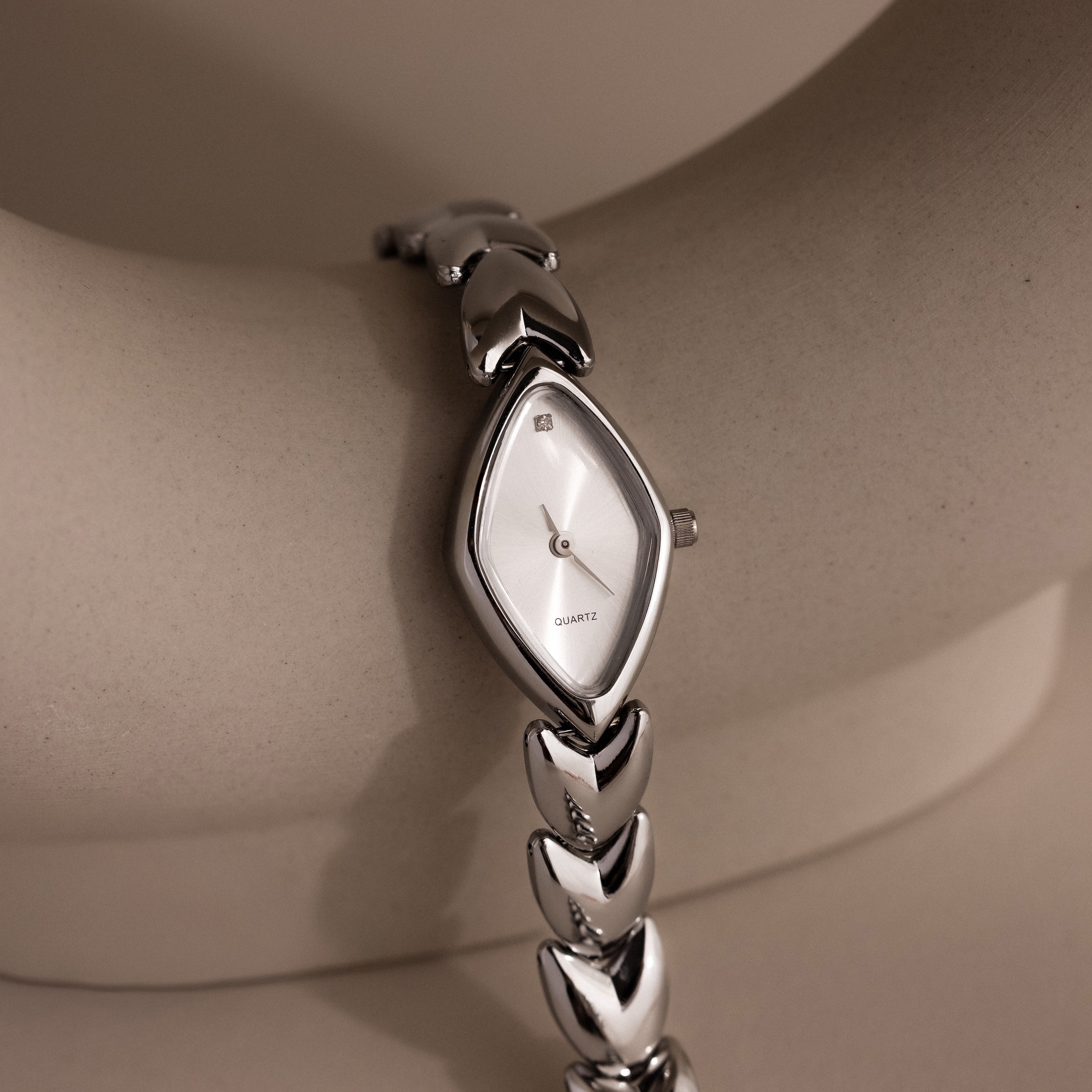 Angled view of a silver wristwatch with a marquise-shaped dial, resting against a neutral ceramic arc, showcasing the dial's shimmer and streamlined bracelet.