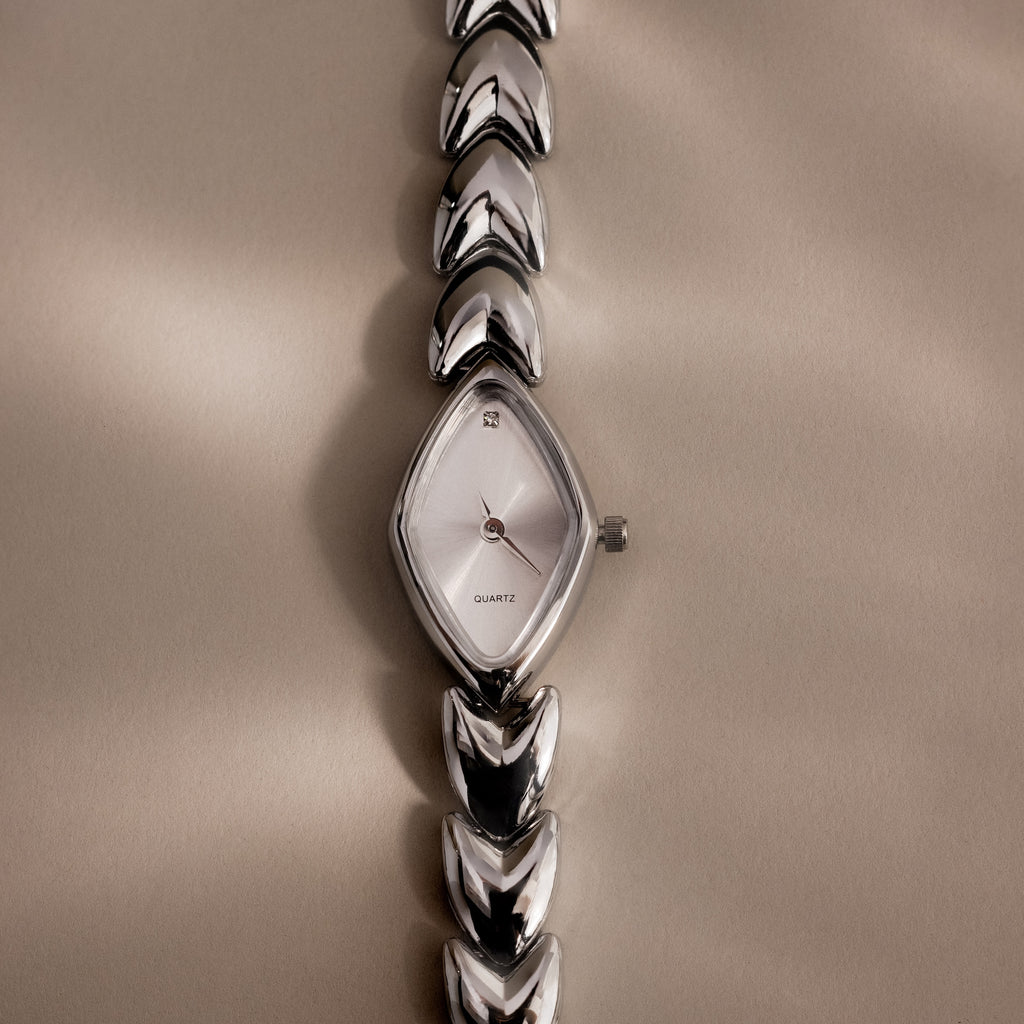 Overhead product shot of a silver geometric oval watch laid flat on a soft beige background, emphasizing its sharp reflective face and arrow-like band links.
