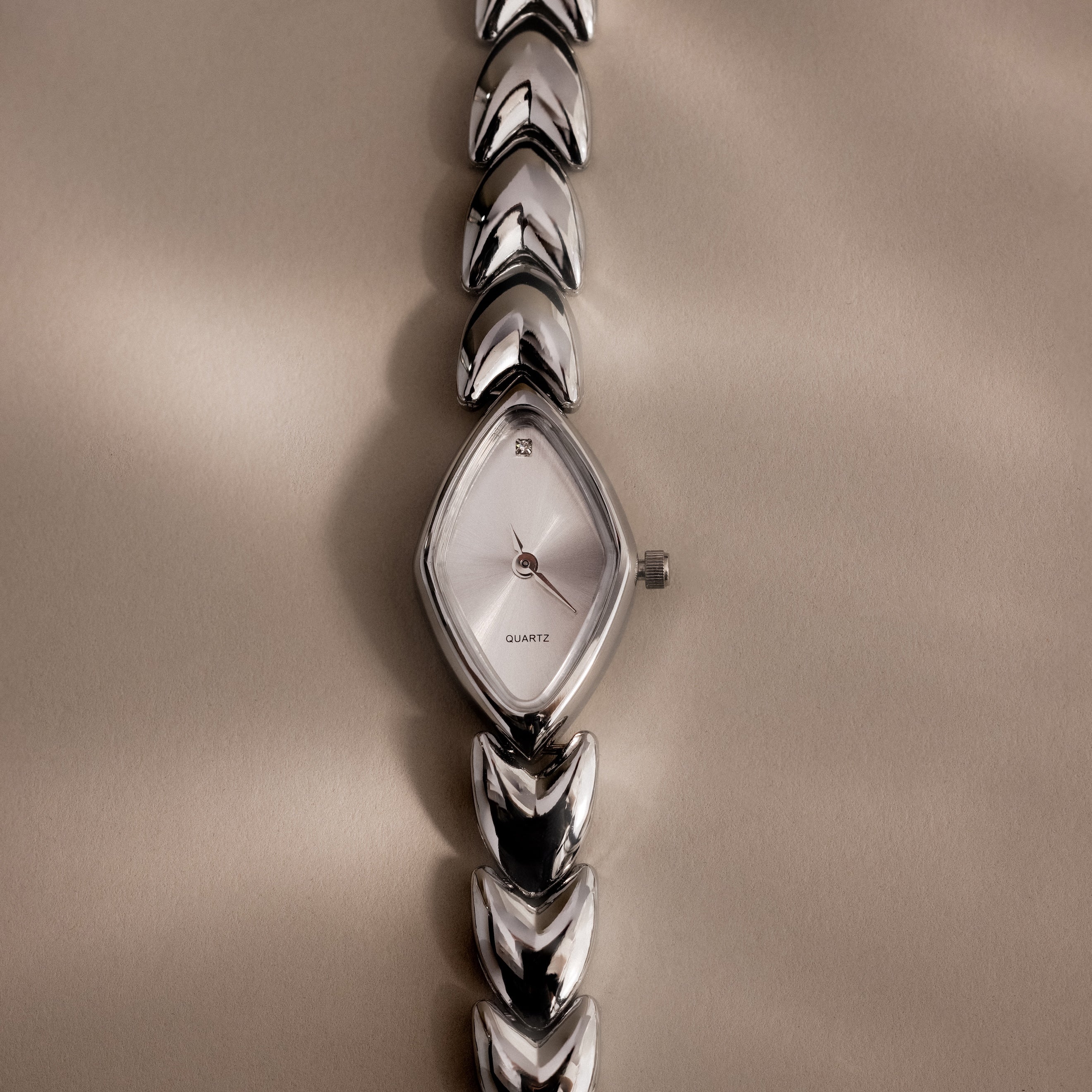 Overhead product shot of a silver geometric oval watch laid flat on a soft beige background, emphasizing its sharp reflective face and arrow-like band links.