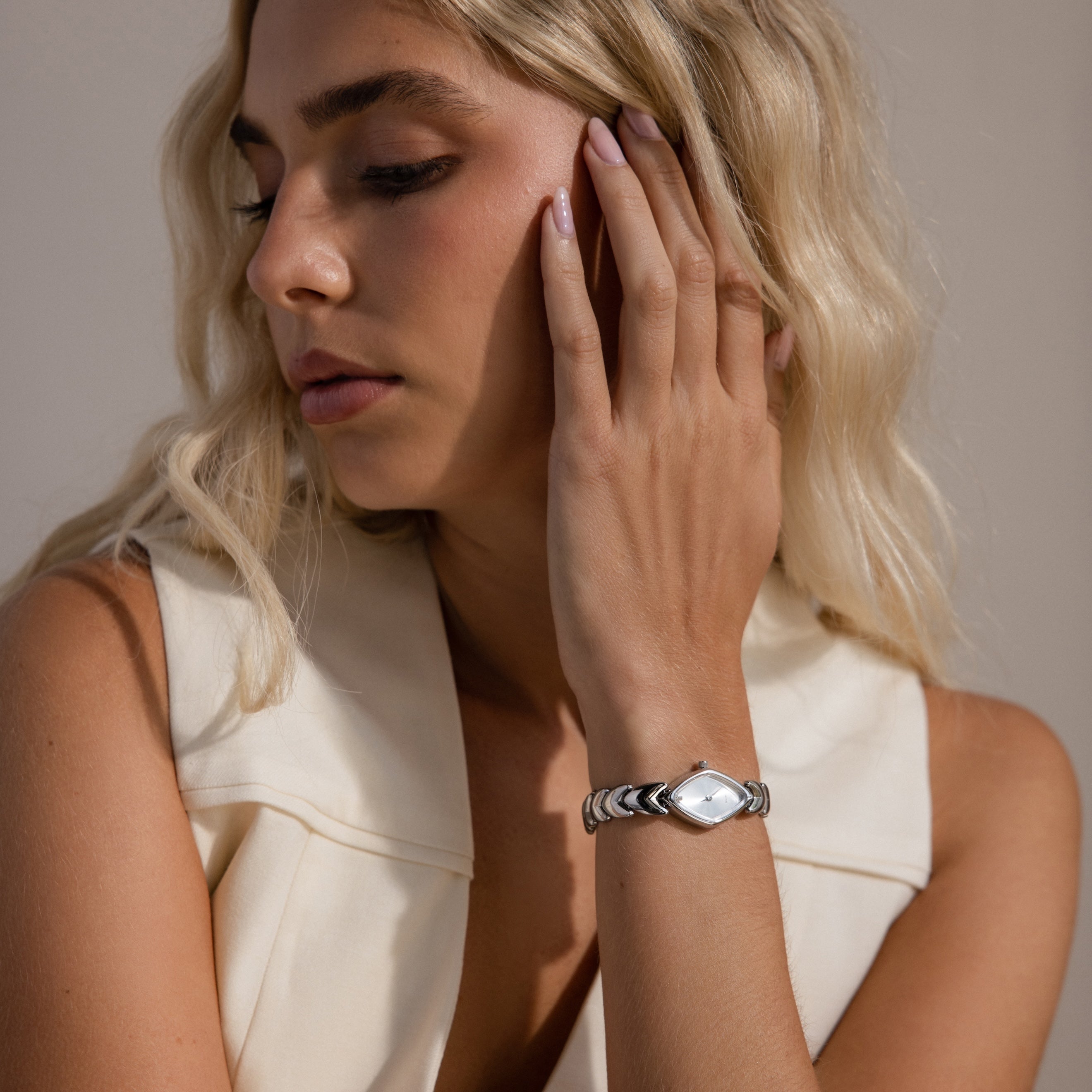 Close-up of a woman with wavy blonde hair wearing a sleeveless cream top and showcasing a silver geometric oval wristwatch with a polished metallic strap.