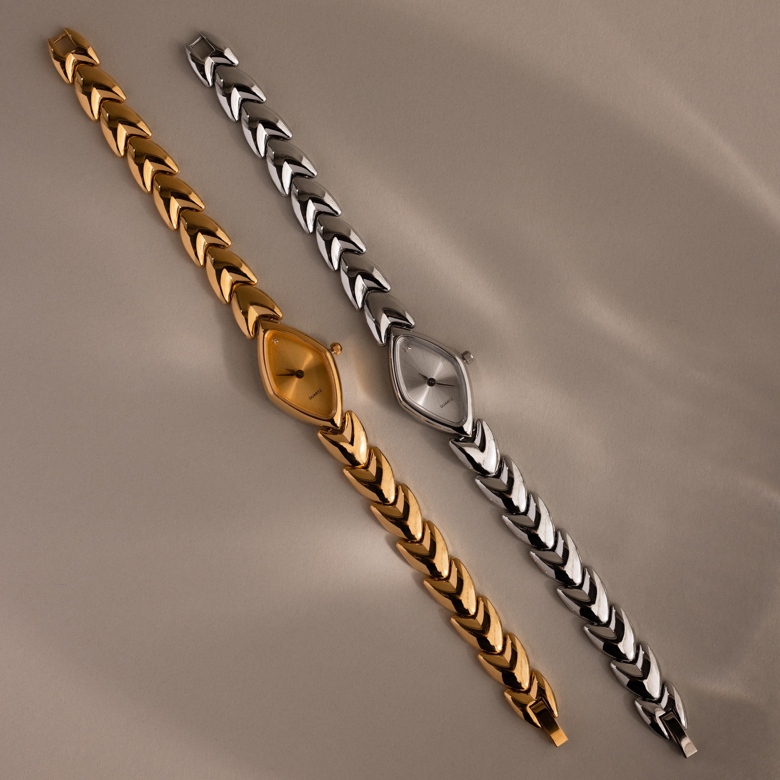 Gold and silver geometric oval watches with chevron-link bands on a neutral background.