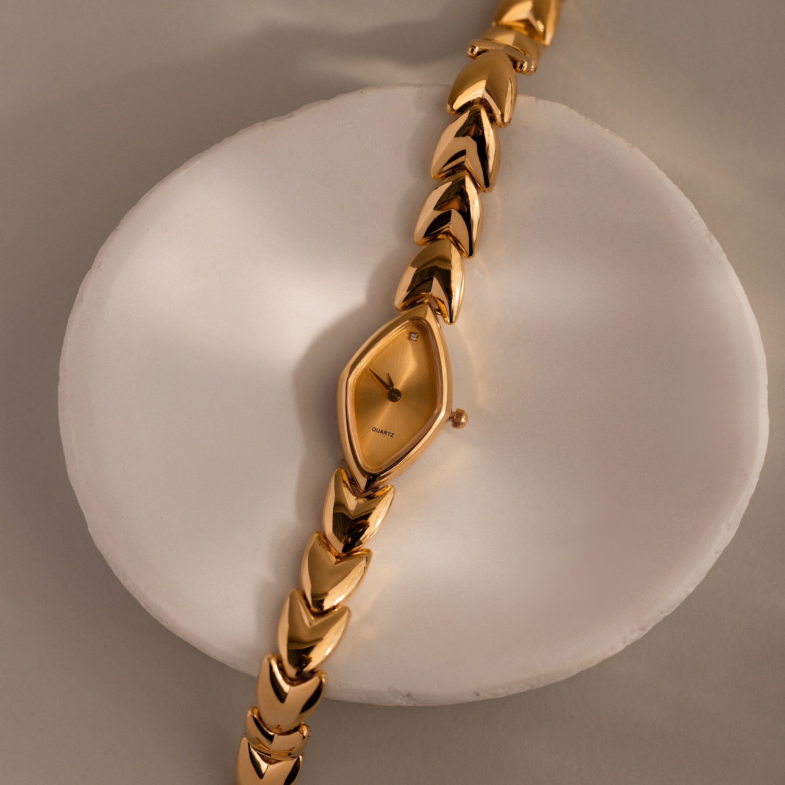 Top-down product shot of the watch laid across a ceramic ring, emphasizing the geometric marquise-shaped face and gold chevron links.
