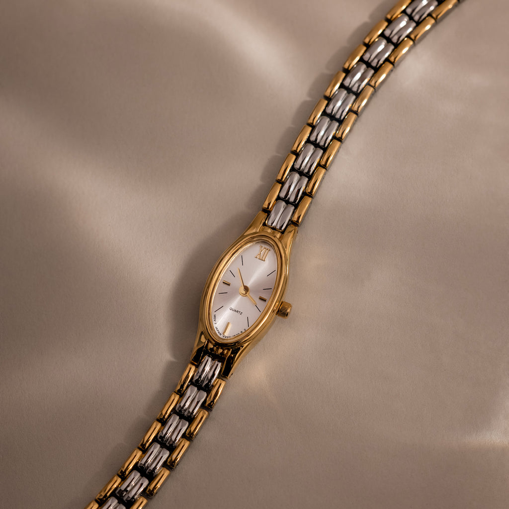 The Slim Oval Timepiece, featuring a polished gold and silver link bracelet, is elegantly displayed on a beige satin surface.