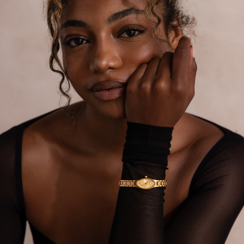 Woman with curly hair wearing a sheer black top and the Slim Oval Timepiece in 18K Gold, its polished link bracelet visible as she rests her chin on her hand.