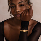 Woman with curly hair wearing a sheer black top and the Slim Oval Timepiece in 18K Gold, its polished link bracelet visible as she rests her chin on her hand.