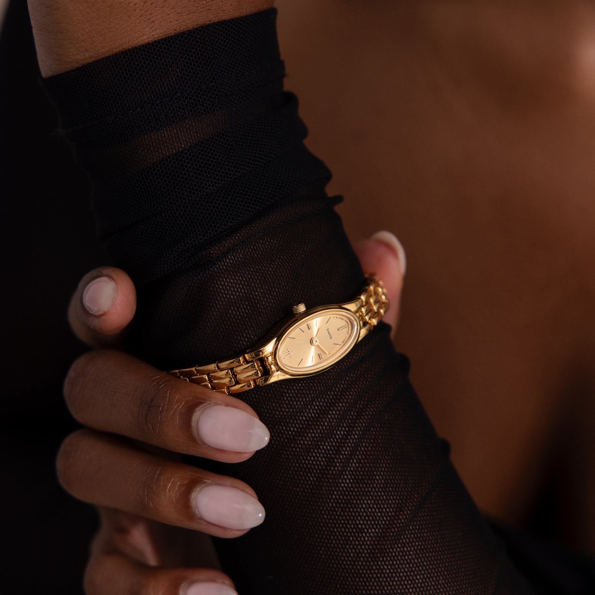 A manicured hand wears the Slim Oval Timepiece, featuring a gold polished link bracelet, over a black sheer glove.