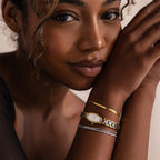 A woman with curly hair rests her head on her arm, wearing the Slim Oval Timepiece in Mixed Metal and three delicate bracelets, including a polished link bracelet.