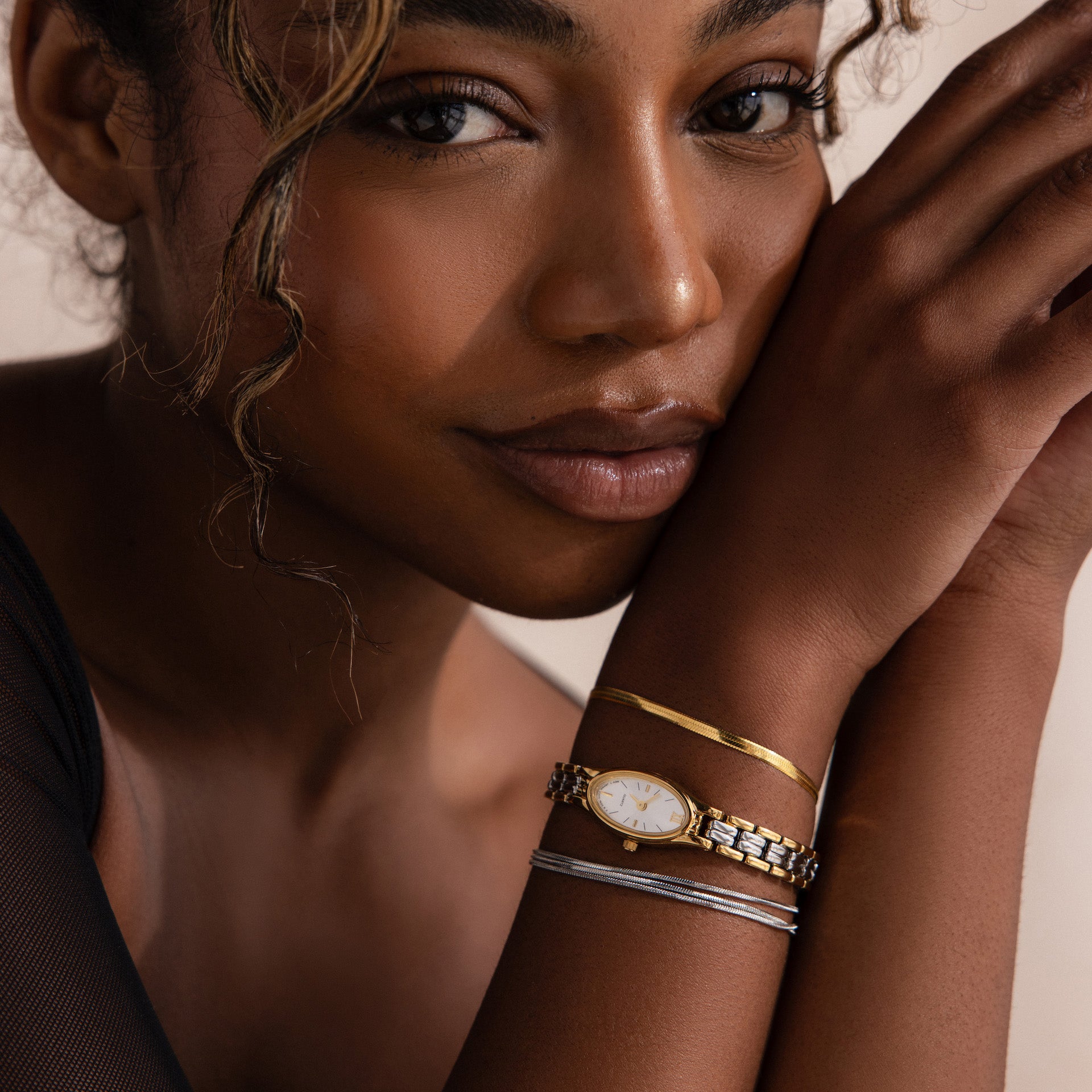 A woman with curly hair rests her head on her arm, wearing the Slim Oval Timepiece in Mixed Metal and three delicate bracelets, including a polished link bracelet.