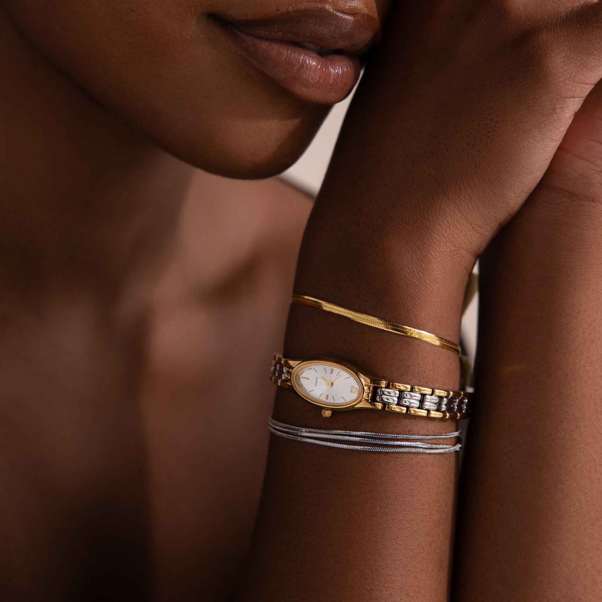 A close-up of a woman's wrist showcases the Slim Oval Timepiece and a polished link bracelet as she thoughtfully rests her chin on her hand.