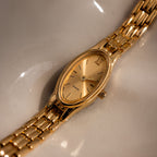 The Slim Oval Timepiece in 18K Gold, featuring a polished link bracelet, is showcased on a smooth, light-colored surface for a modern minimalist look.