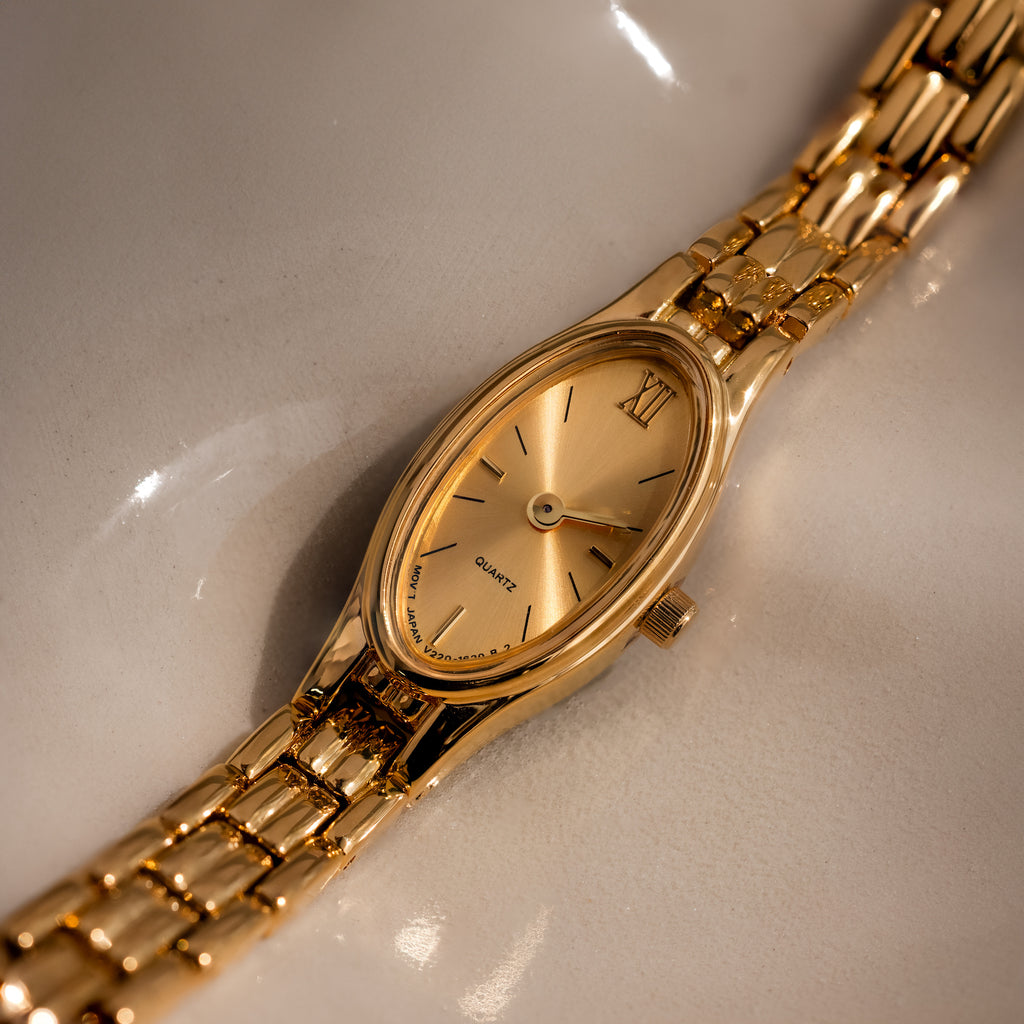 The Slim Oval Timepiece showcases a gold oval face and a polished link bracelet, elegantly presented on a reflective cream surface.