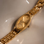 The Slim Oval Timepiece showcases a gold oval face and a polished link bracelet, elegantly presented on a reflective cream surface.