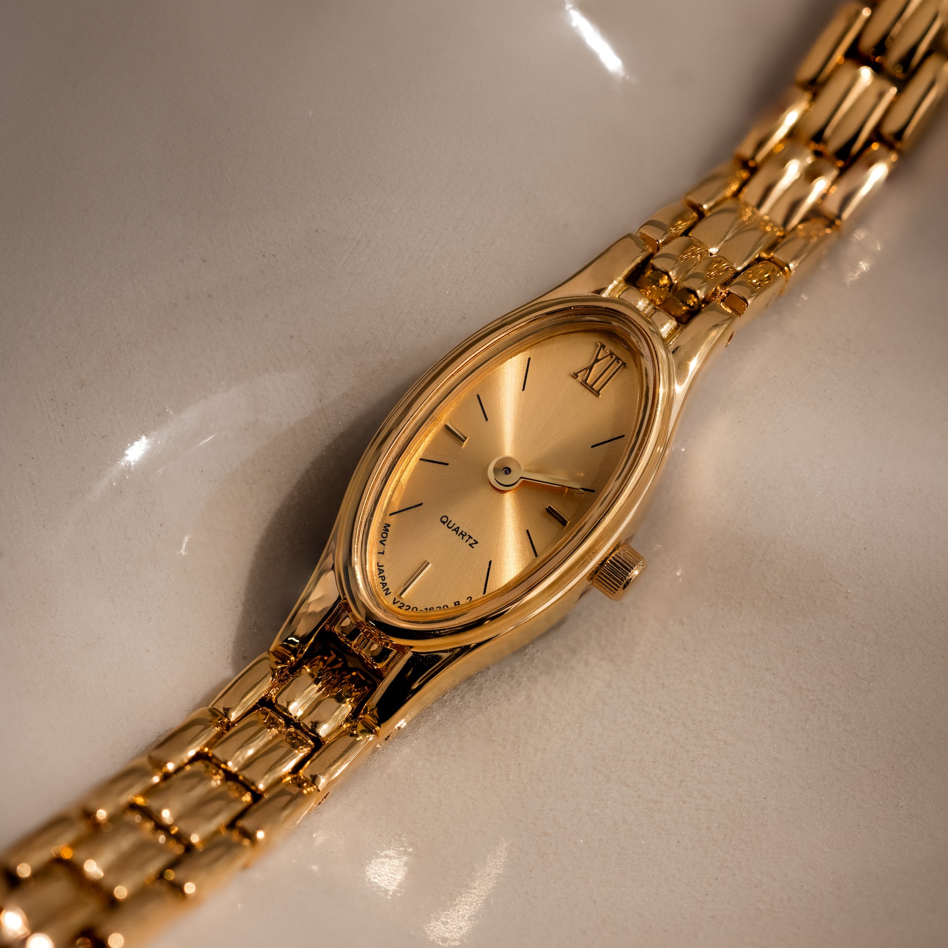 The Slim Oval Timepiece showcases a gold oval face and a polished link bracelet, elegantly presented on a reflective cream surface.