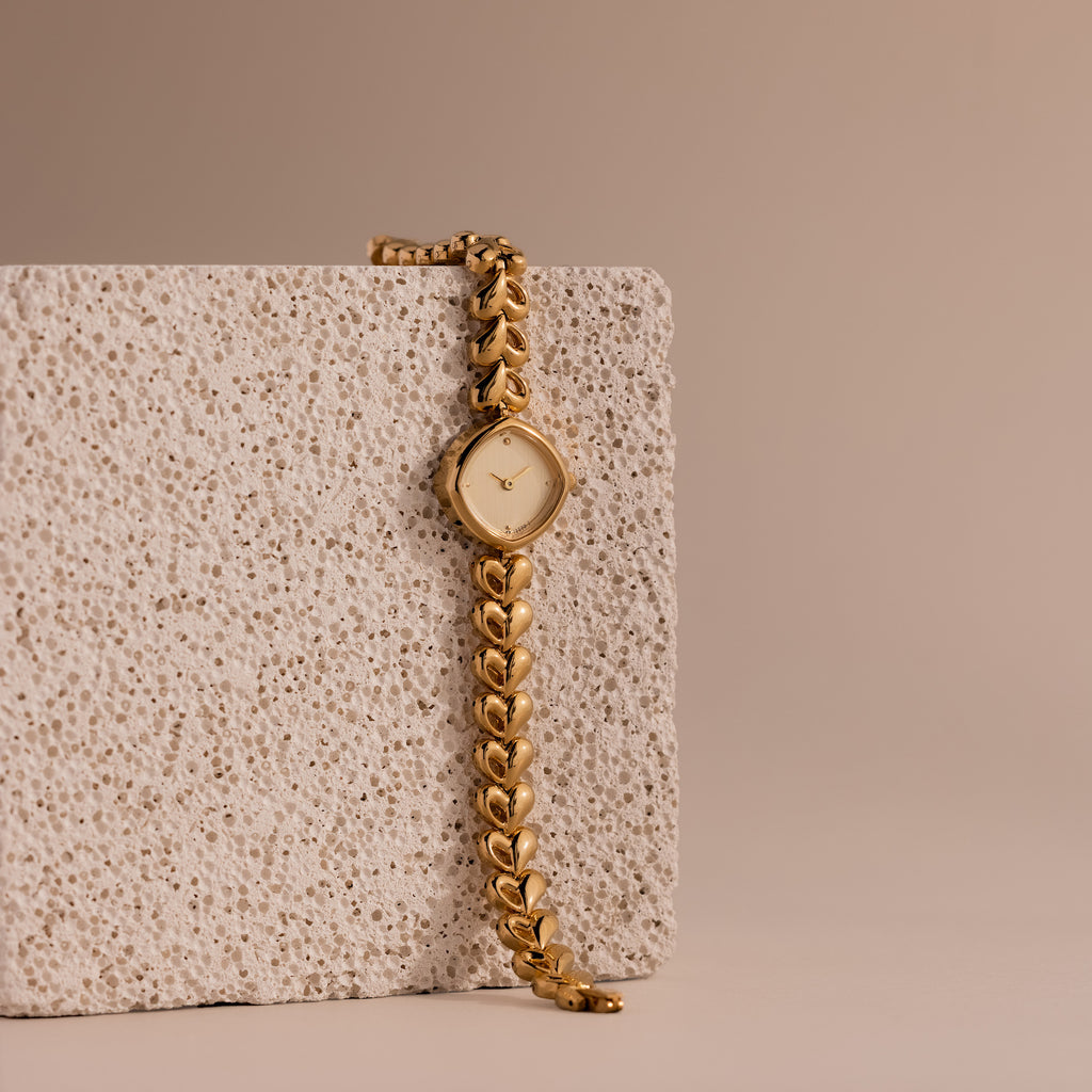 The Heart Link Timepiece in 18K Gold, featuring a minimalist dial and heart-shaped links, rests against a textured stone block on a beige background.