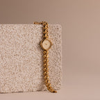 The Heart Link Timepiece in 18K Gold, featuring a minimalist dial and heart-shaped links, rests against a textured stone block on a beige background.