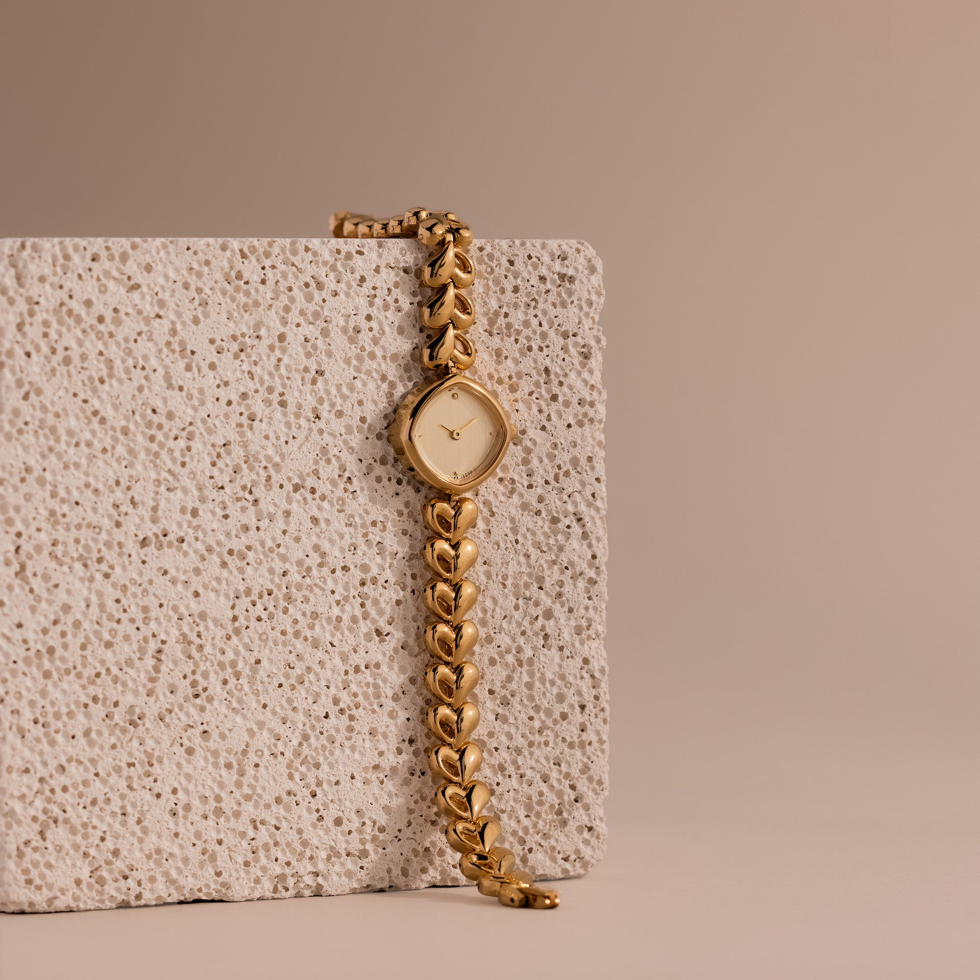The Heart Link Timepiece in 18K Gold, featuring a minimalist dial and heart-shaped links, rests against a textured stone block on a beige background.