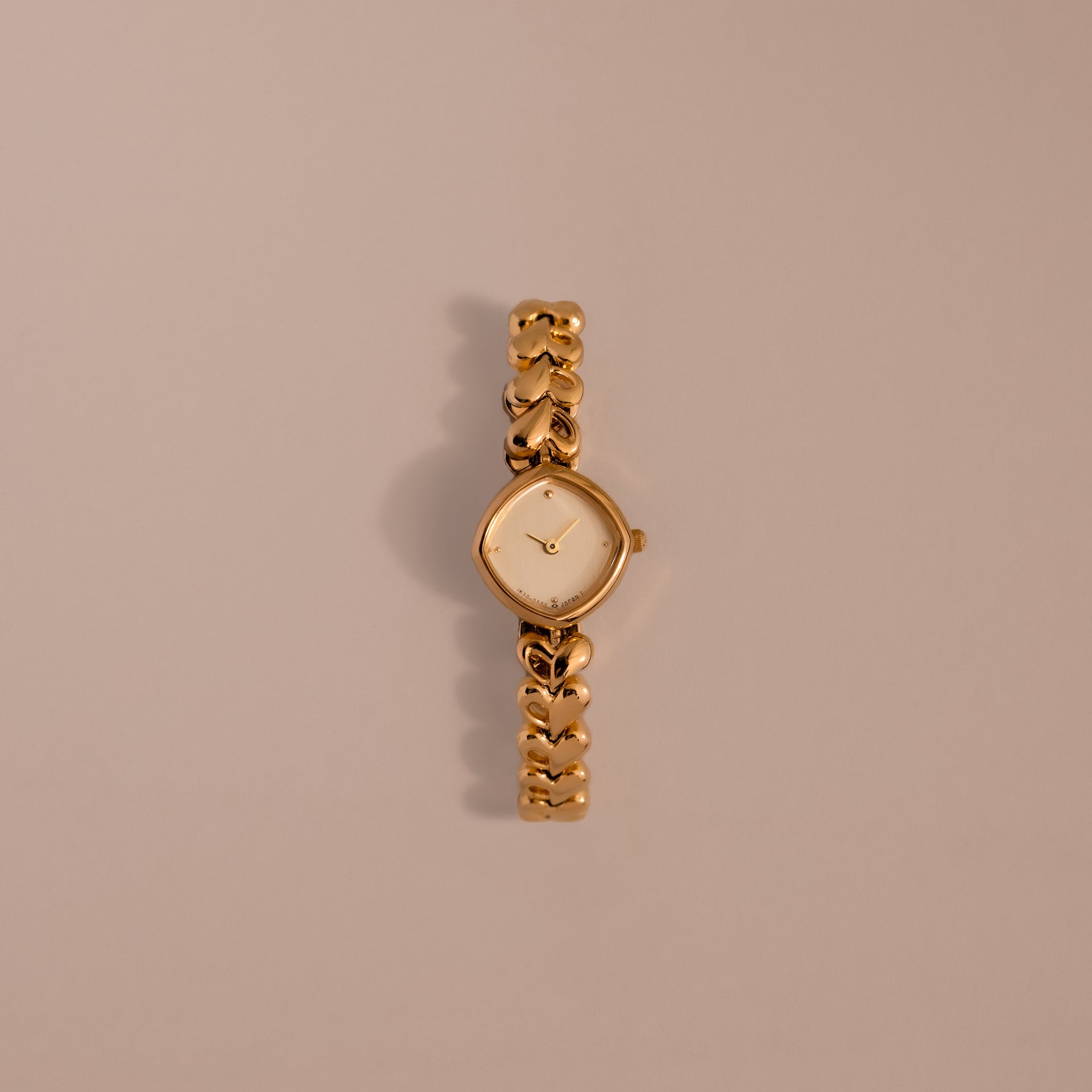 The Heart Link Timepiece is a gold wristwatch featuring heart-shaped links and a minimalist dial, displayed on a light beige background.