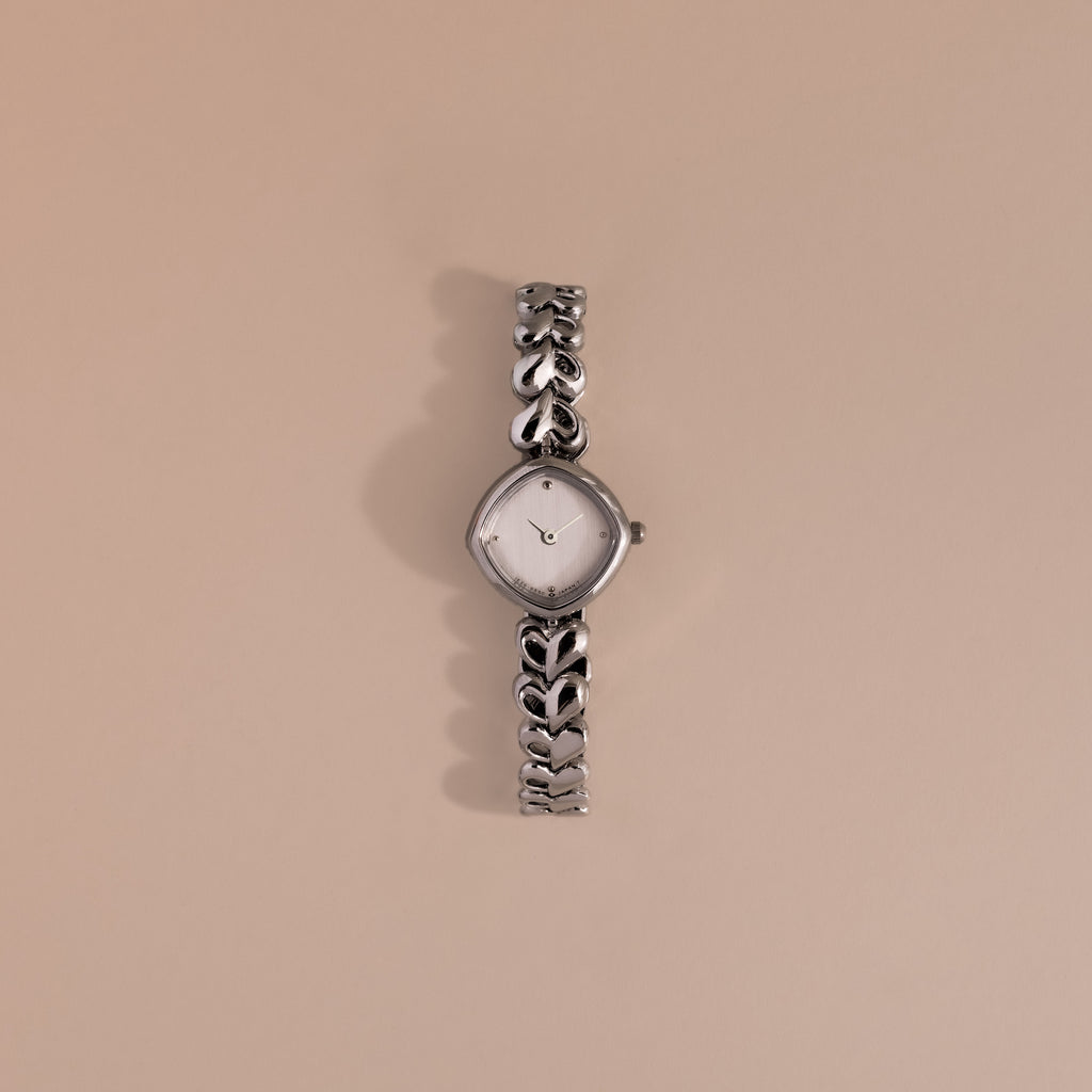 The Heart Link Timepiece in White Gold features heart-shaped links and a minimalist dial, showcased against a simple beige background.