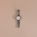 The Heart Link Timepiece in White Gold features heart-shaped links and a minimalist dial, showcased against a simple beige background.