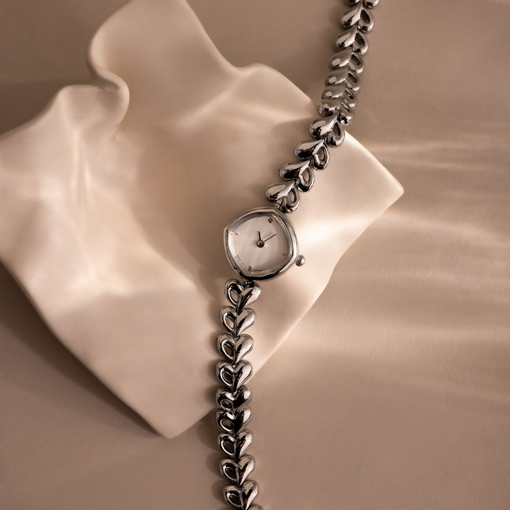 The Heart Link Timepiece, featuring a minimalist dial and heart-shaped links in silver, is displayed on a wavy, cream-colored surface.