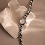 The Heart Link Timepiece, featuring a minimalist dial and heart-shaped links in silver, is displayed on a wavy, cream-colored surface.