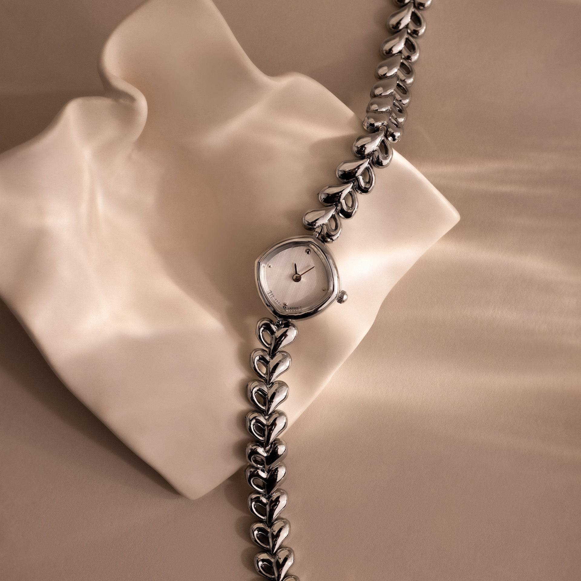 The Heart Link Timepiece features delicate heart-shaped links and a minimalist dial, elegantly displayed on a wavy, cream-colored surface.