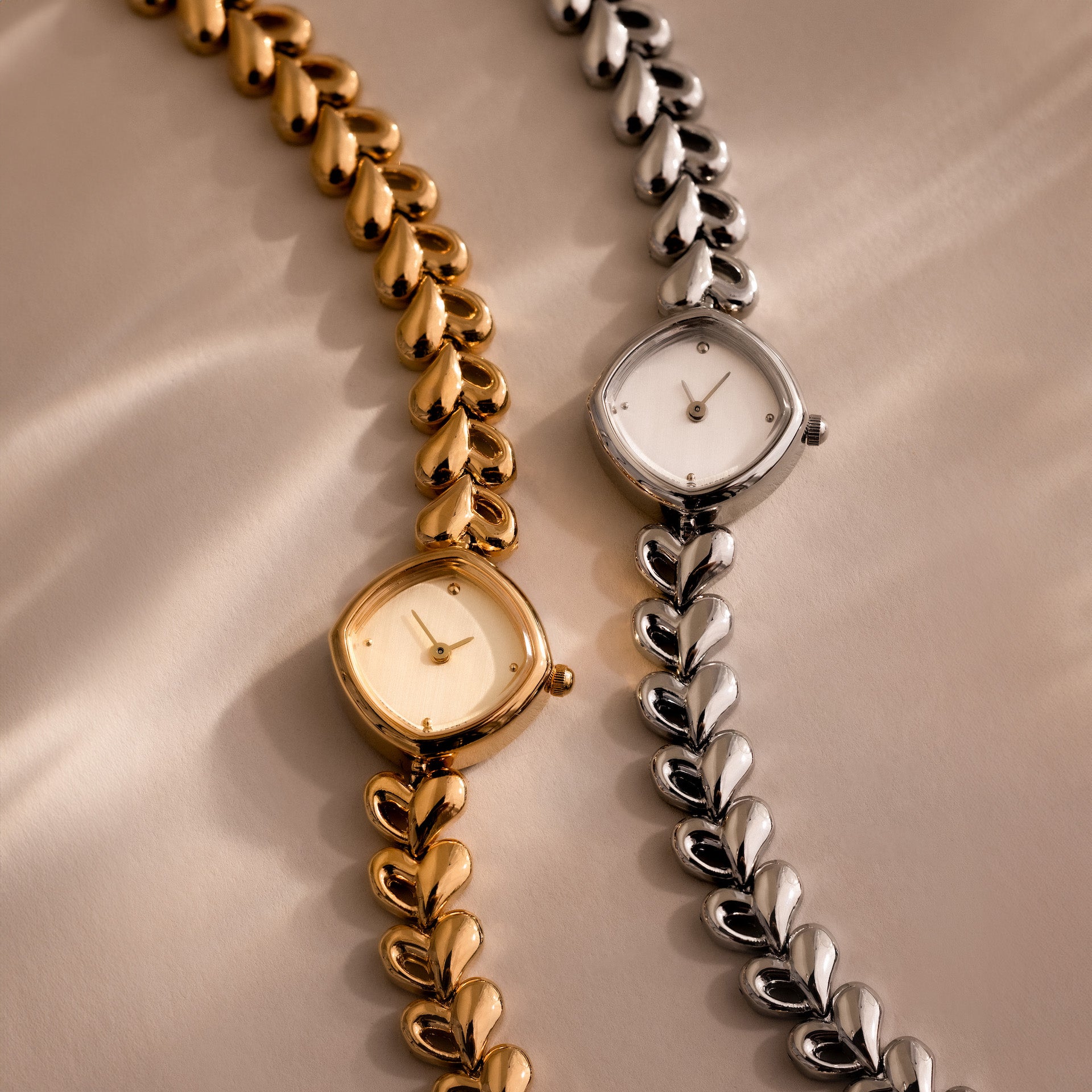 The Heart Link Timepiece, featuring gold and silver designs with heart-shaped links and a minimalist dial, is displayed side by side on a beige surface.