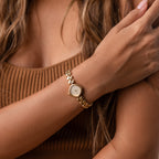 A woman wearing the Heart Link Timepiece, featuring heart-shaped links, crosses her arm over her chest while dressed in a brown ribbed top.