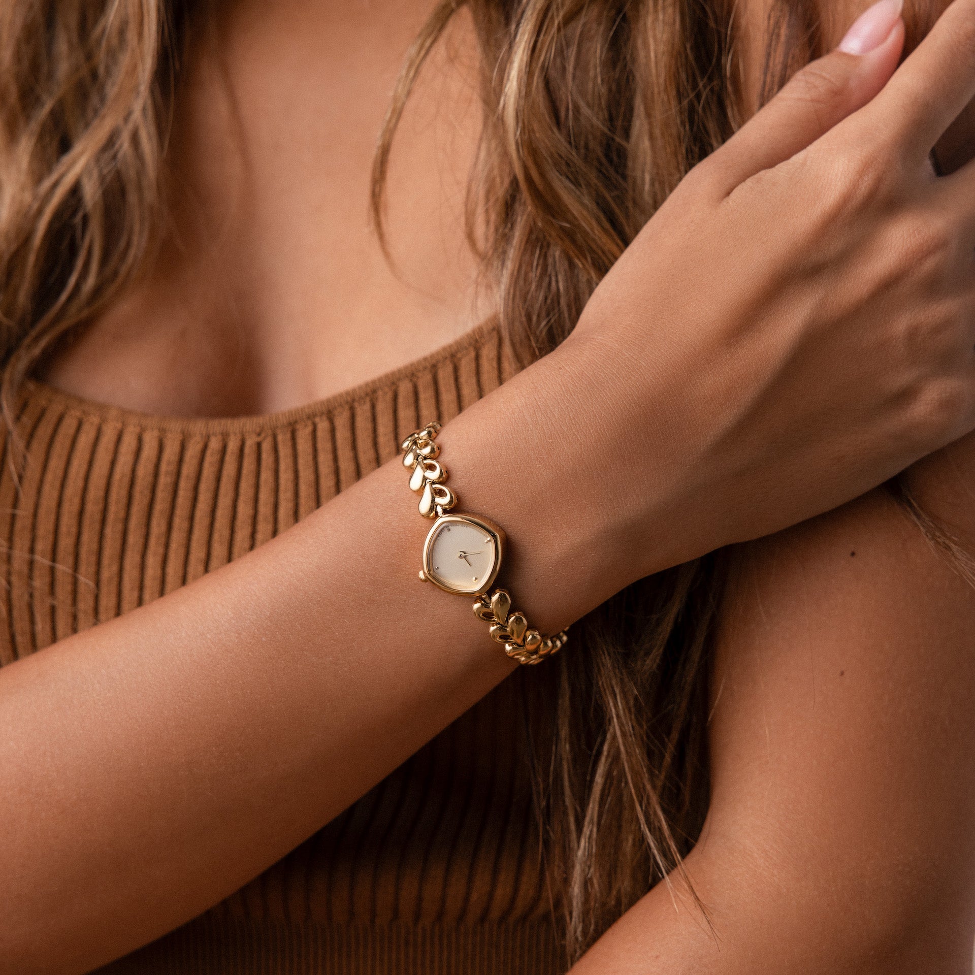 A woman in a brown ribbed tank top wears the Heart Link Timepiece, showcasing heart-shaped links and a leaf-patterned band.