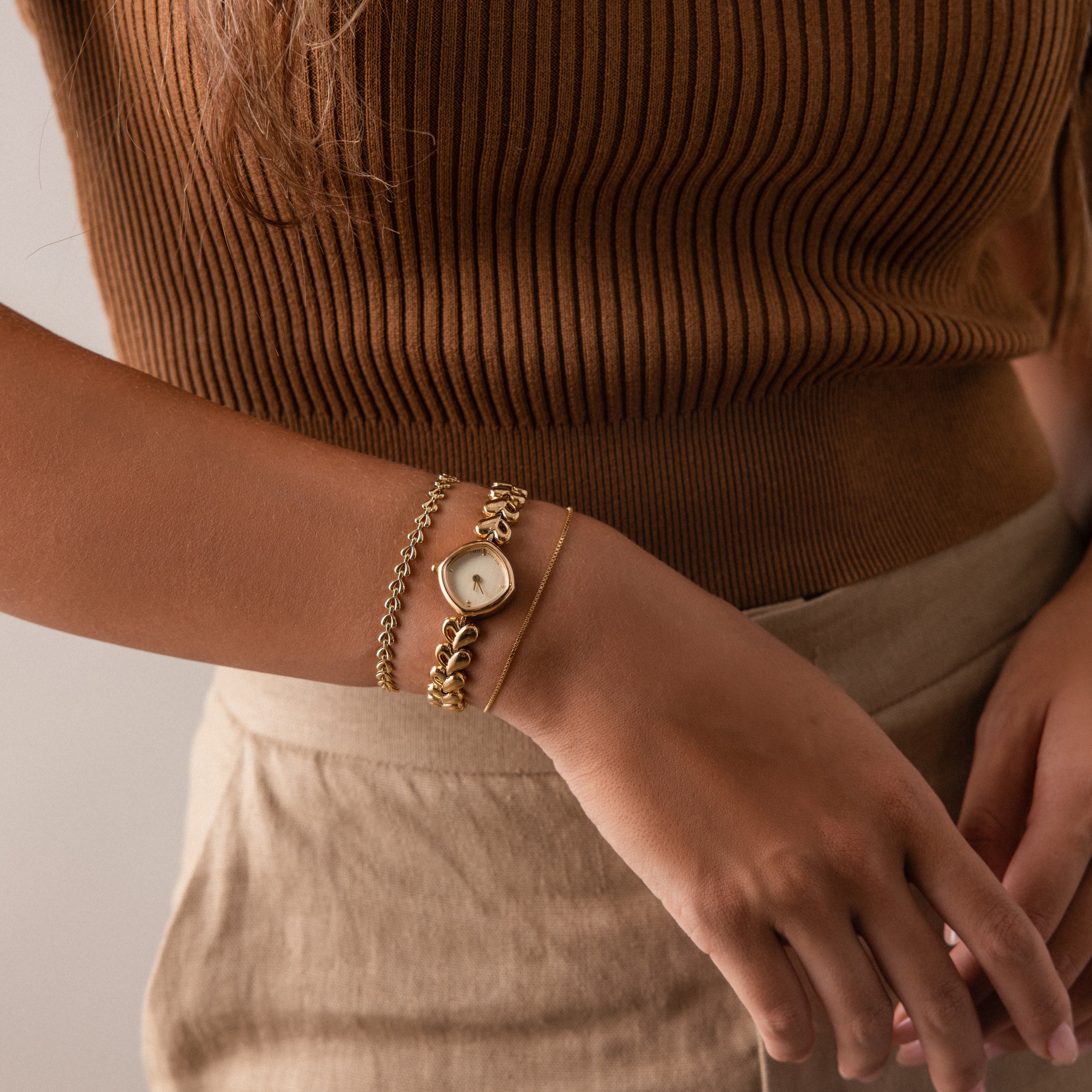 A woman dressed in a brown ribbed top and beige pants accessorizes with curated jewelry, featuring gold bracelets, a watch, and items from the Corporate Gift Box.